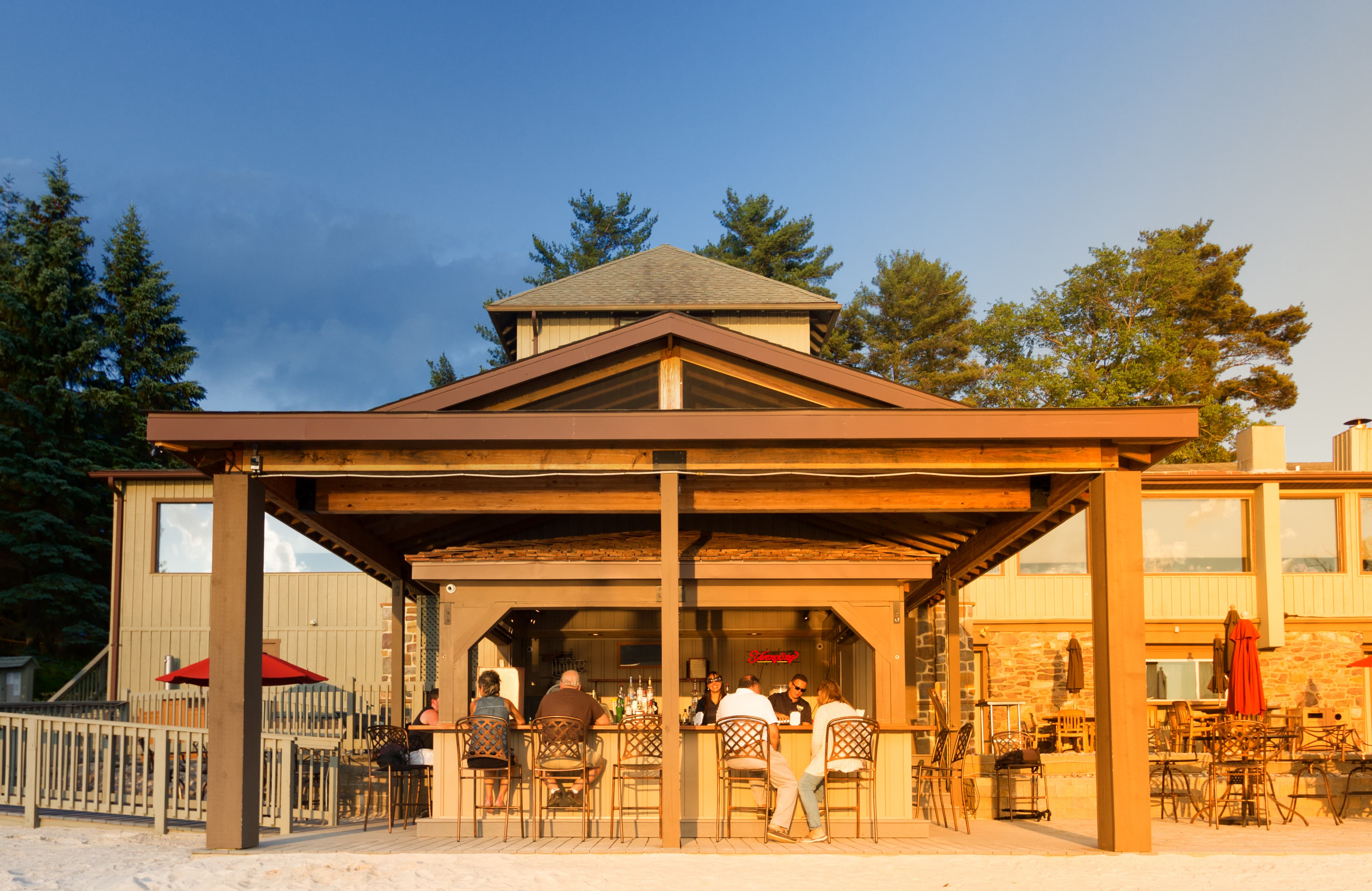 A waterfront bar with patrons enjoying drinks under a shaded structure at sunset.