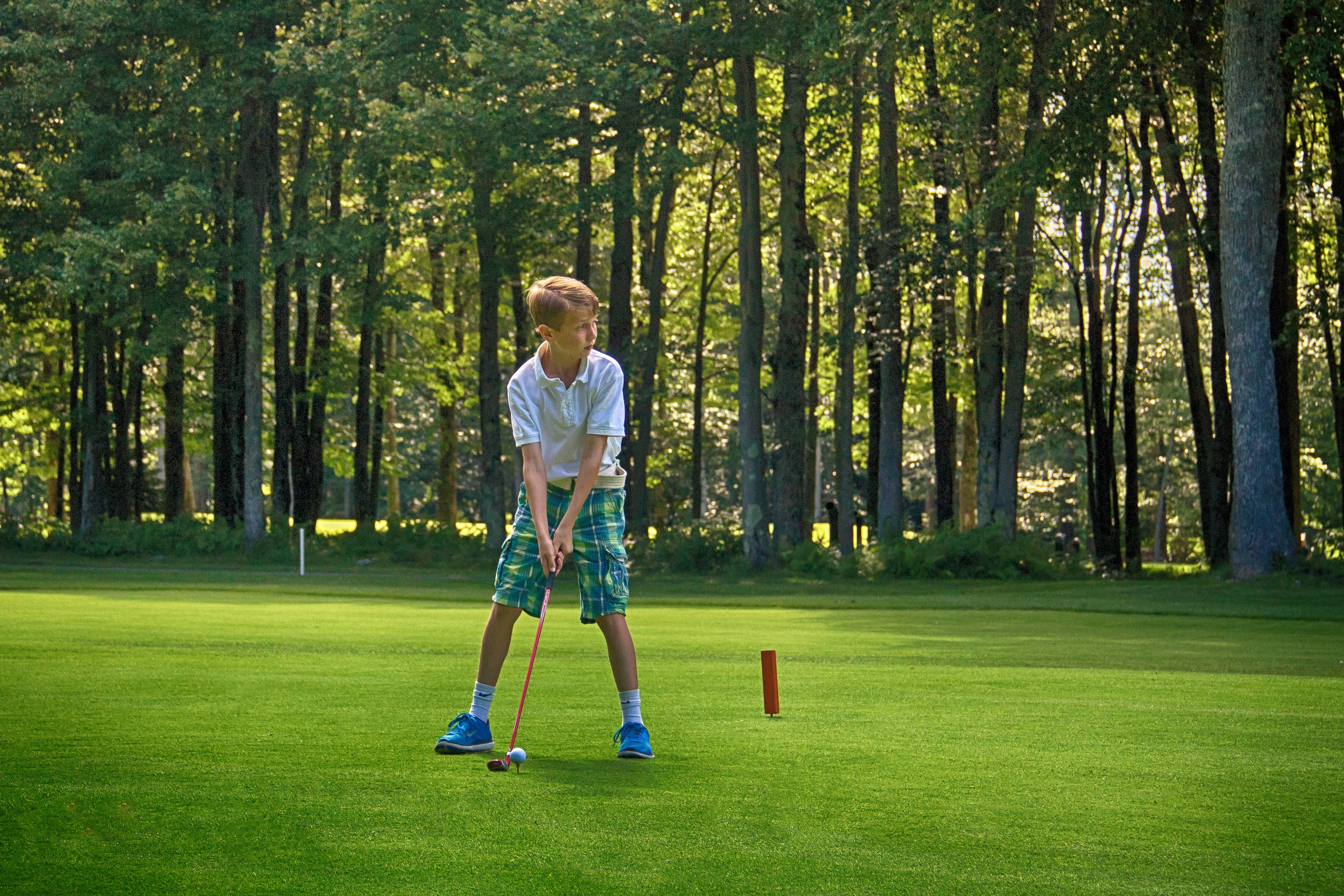A boy prepares to hit a golf ball on a lush green course surrounded by trees.