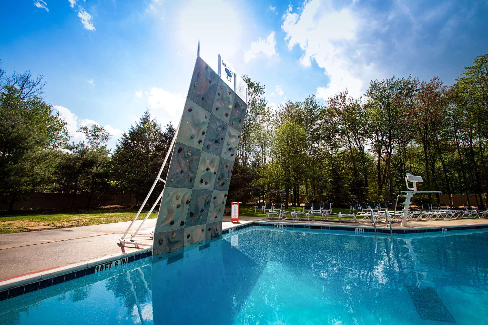 A climbing wall stands next to a swimming pool surrounded by trees and lounge chairs.