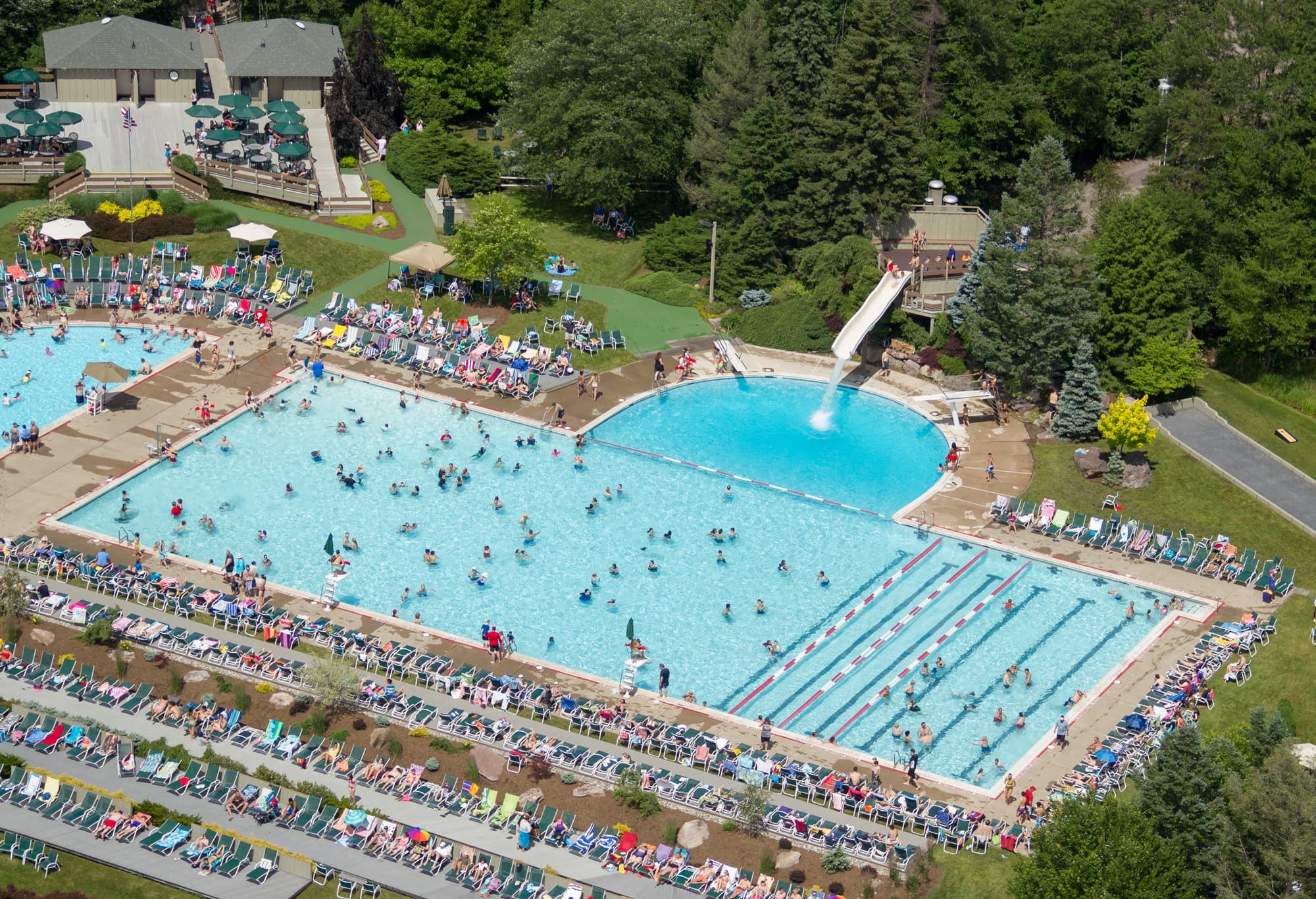 Aerial view of a crowded recreational pool area with multiple pools, a waterslide, and sunbathers lounging nearby.