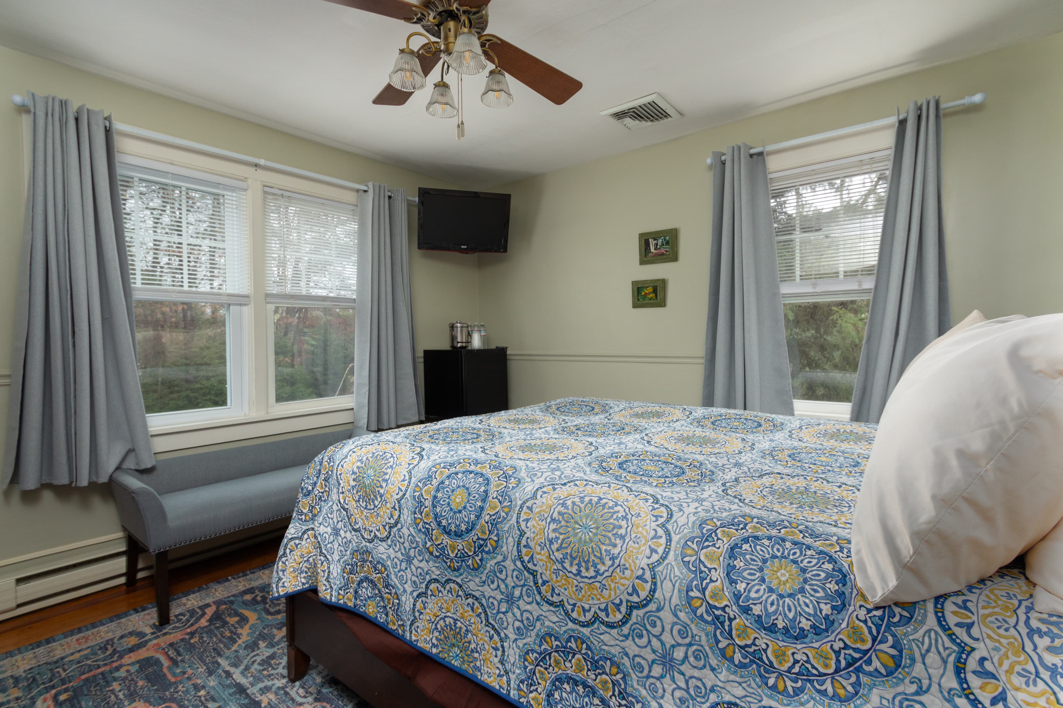 A bright bedroom with light green walls, a queen bed covered in a blue and yellow patterned quilt, and two windows with pale blue curtains.