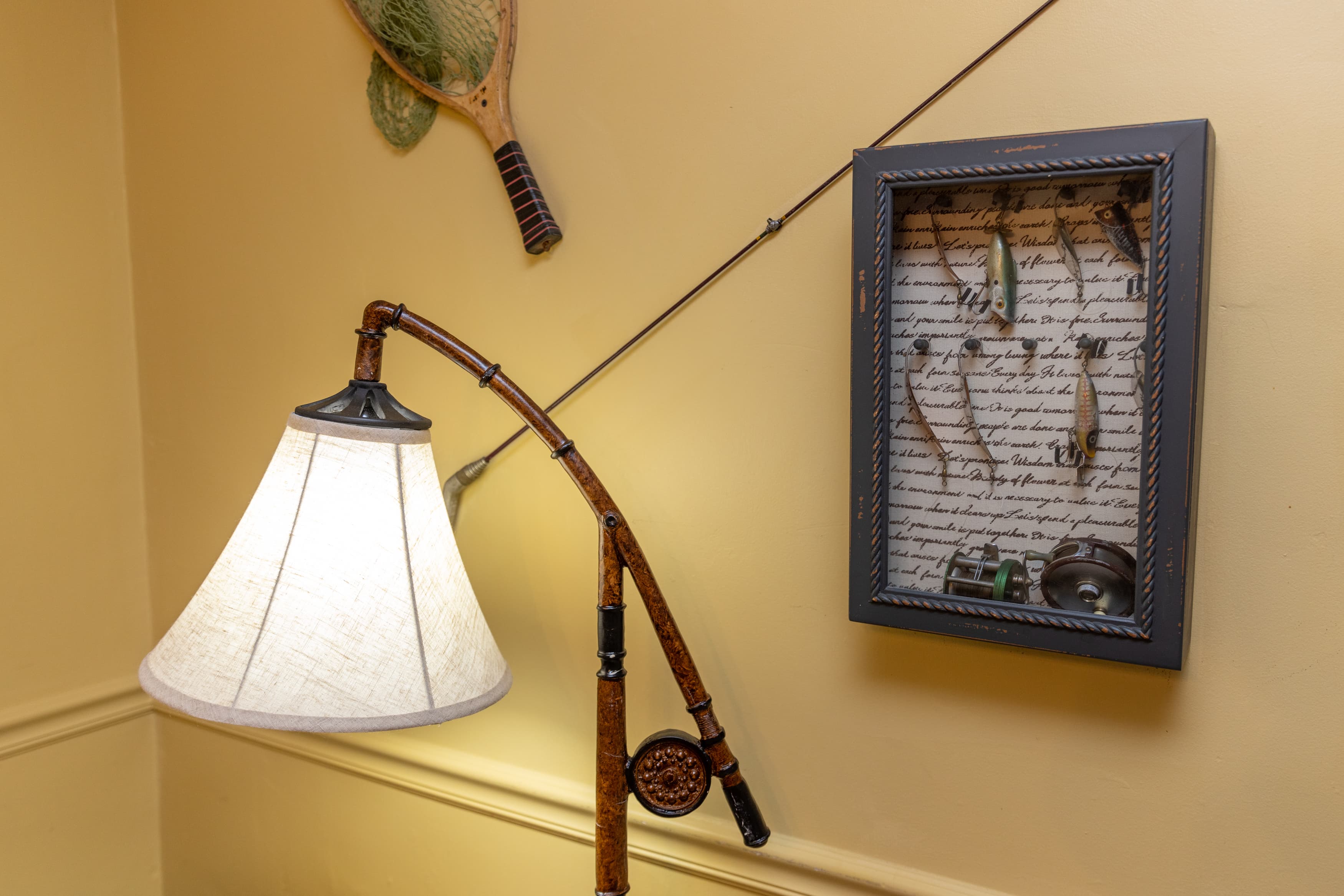A rustic fishing-themed display with a shadowbox of fishing lures, a vintage landing net, and a floor lamp with a curved pole.