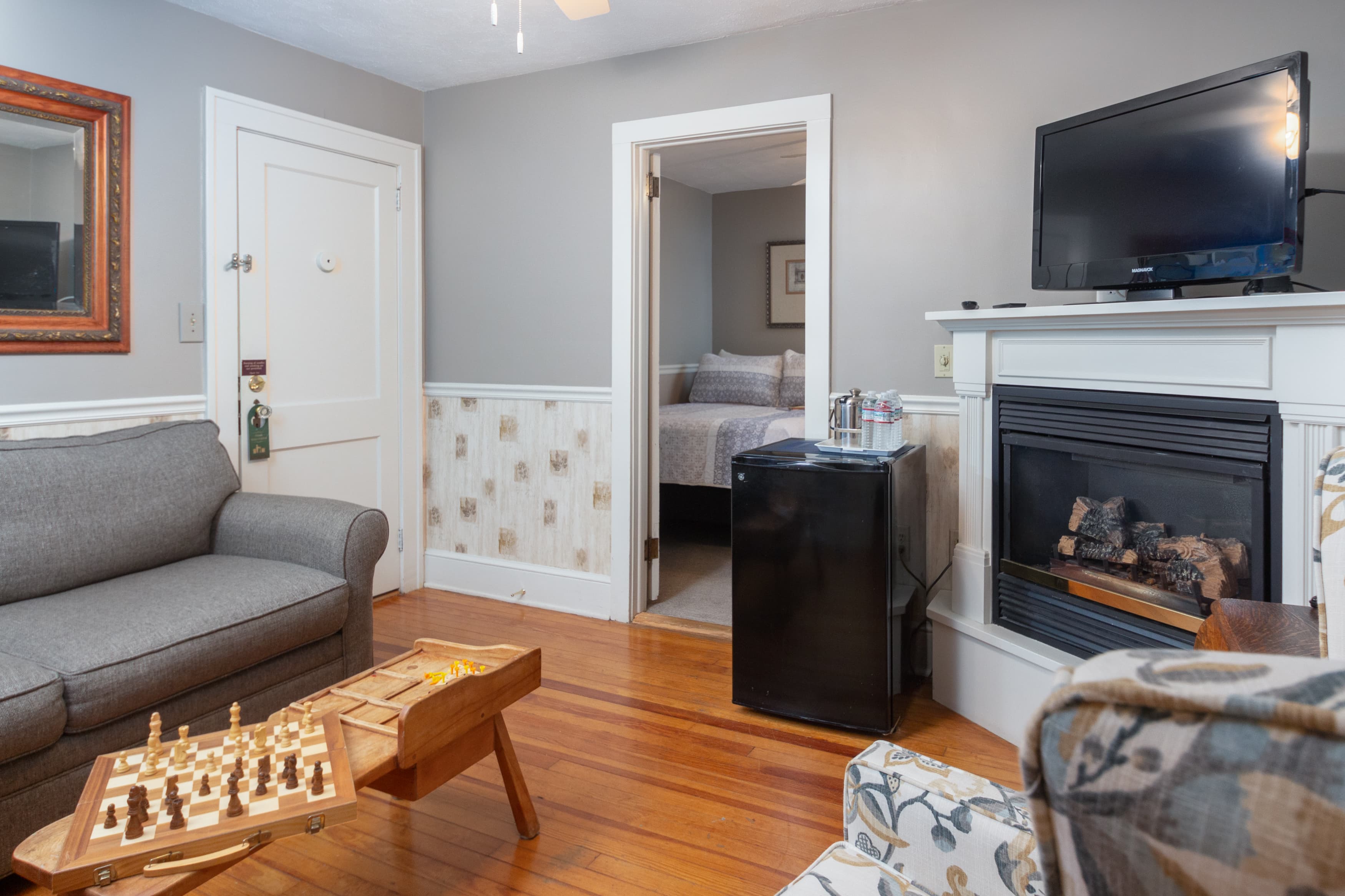 A cozy suite living area with a gray sofa, a small fireplace, hardwood floors, a mini-fridge, and a chess board on the coffee table.