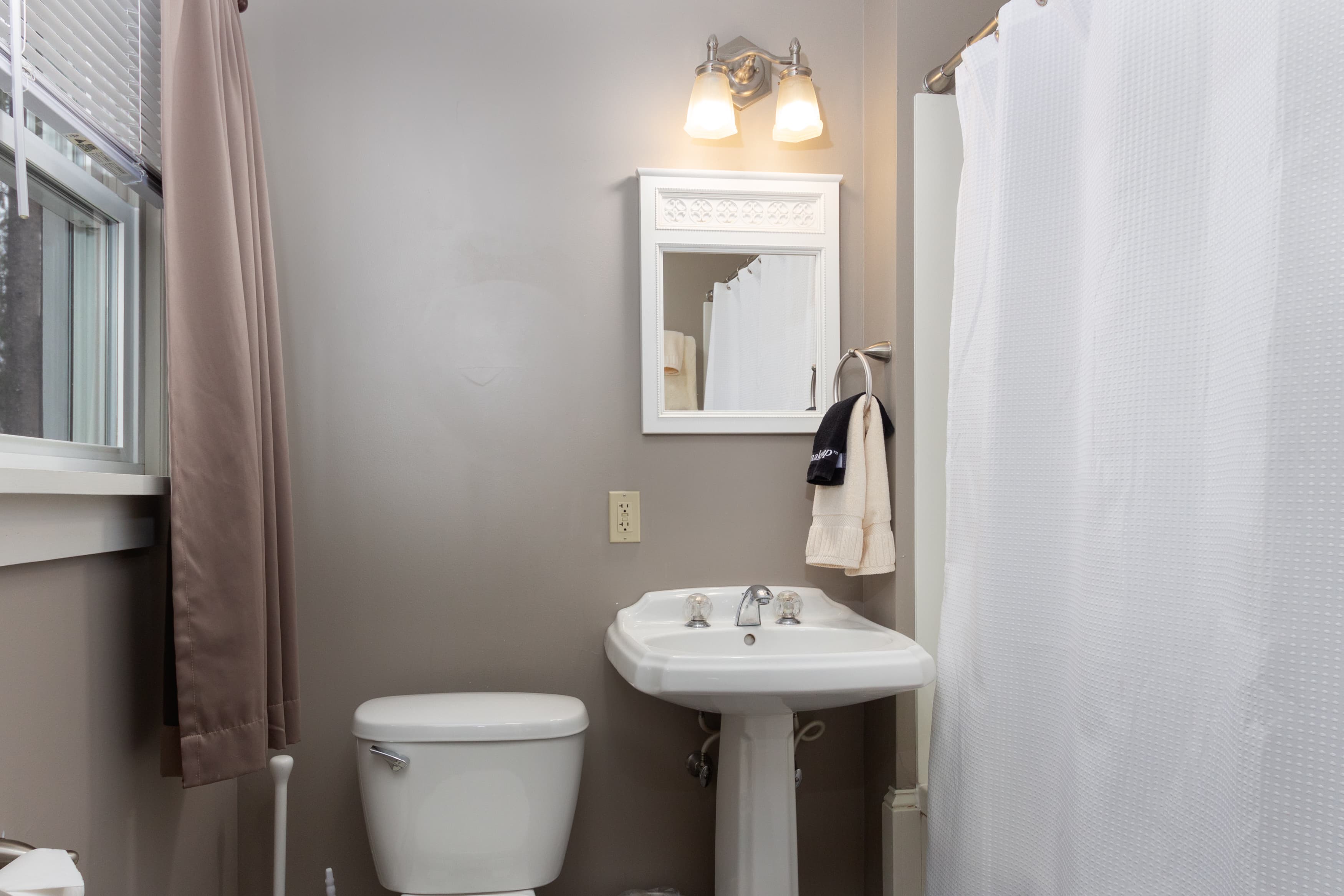 A clean, modern bathroom with gray walls, a white pedestal sink, a toilet, a tub/shower combo, and dark brown floor tiles.
