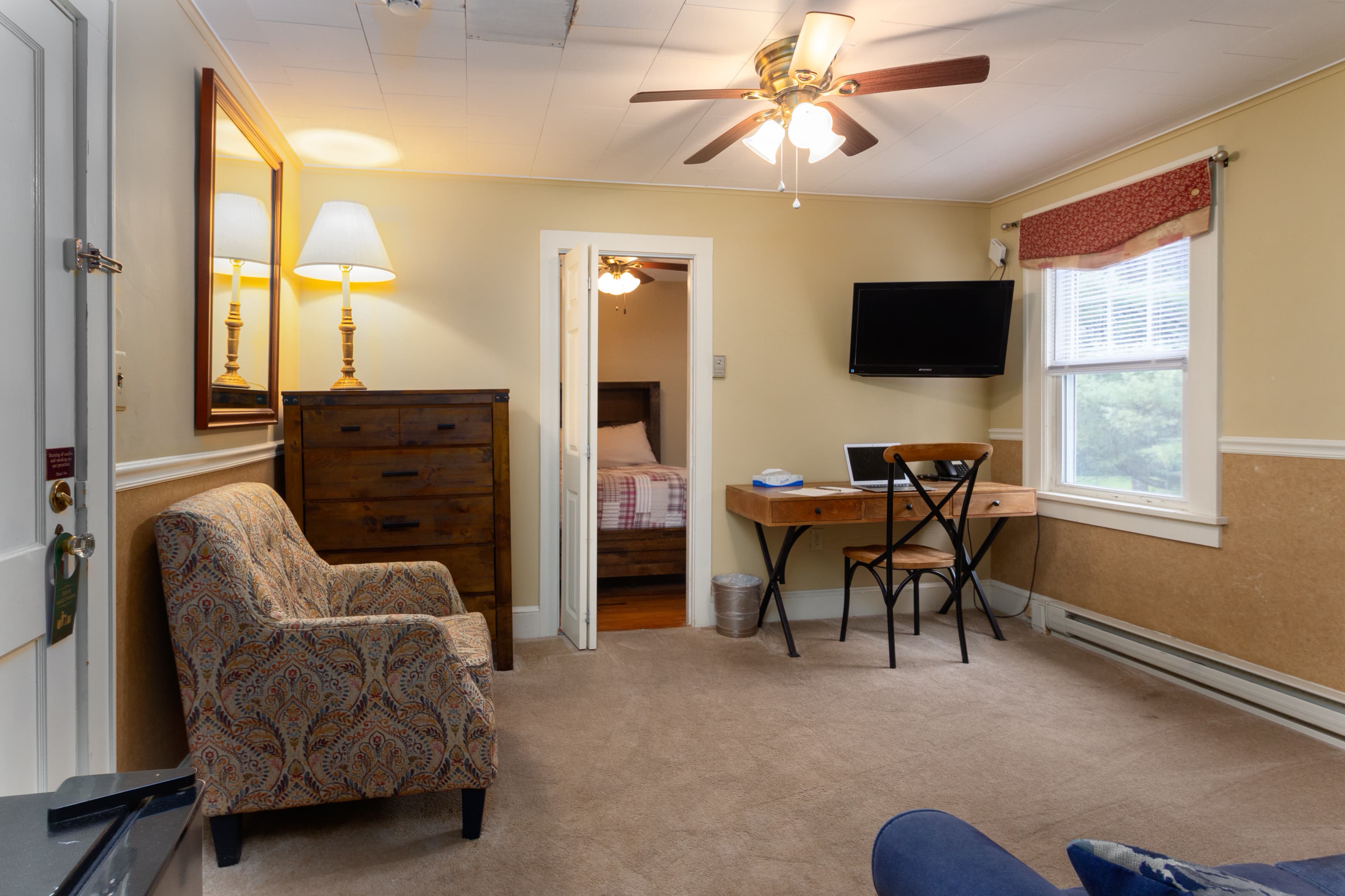 A sitting room with a patterned armchair, a writing desk, and a wall-mounted TV, looking through a doorway into the bedroom.