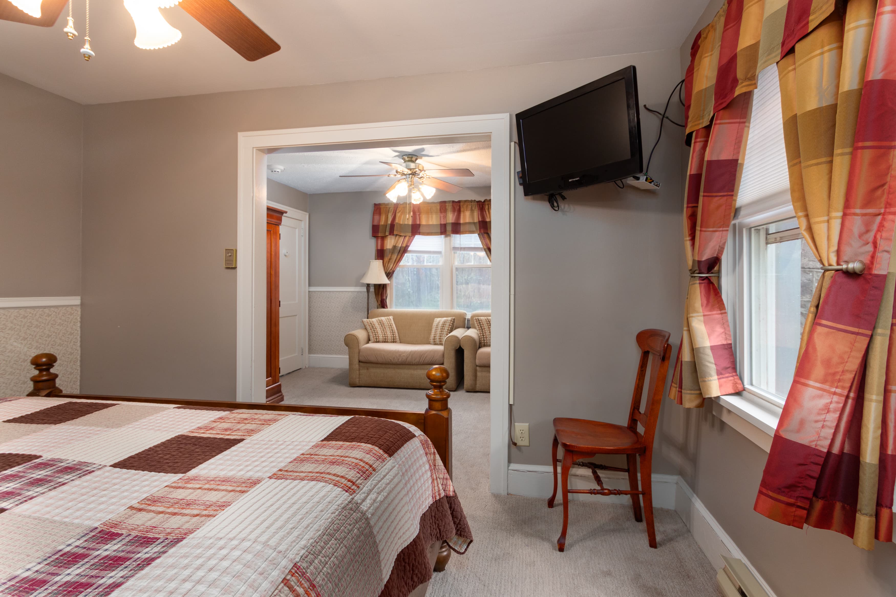 An inviting suite with a plaid quilt on the king bed, a wall-mounted TV, and a view through a doorway into a sitting area with a sofa.