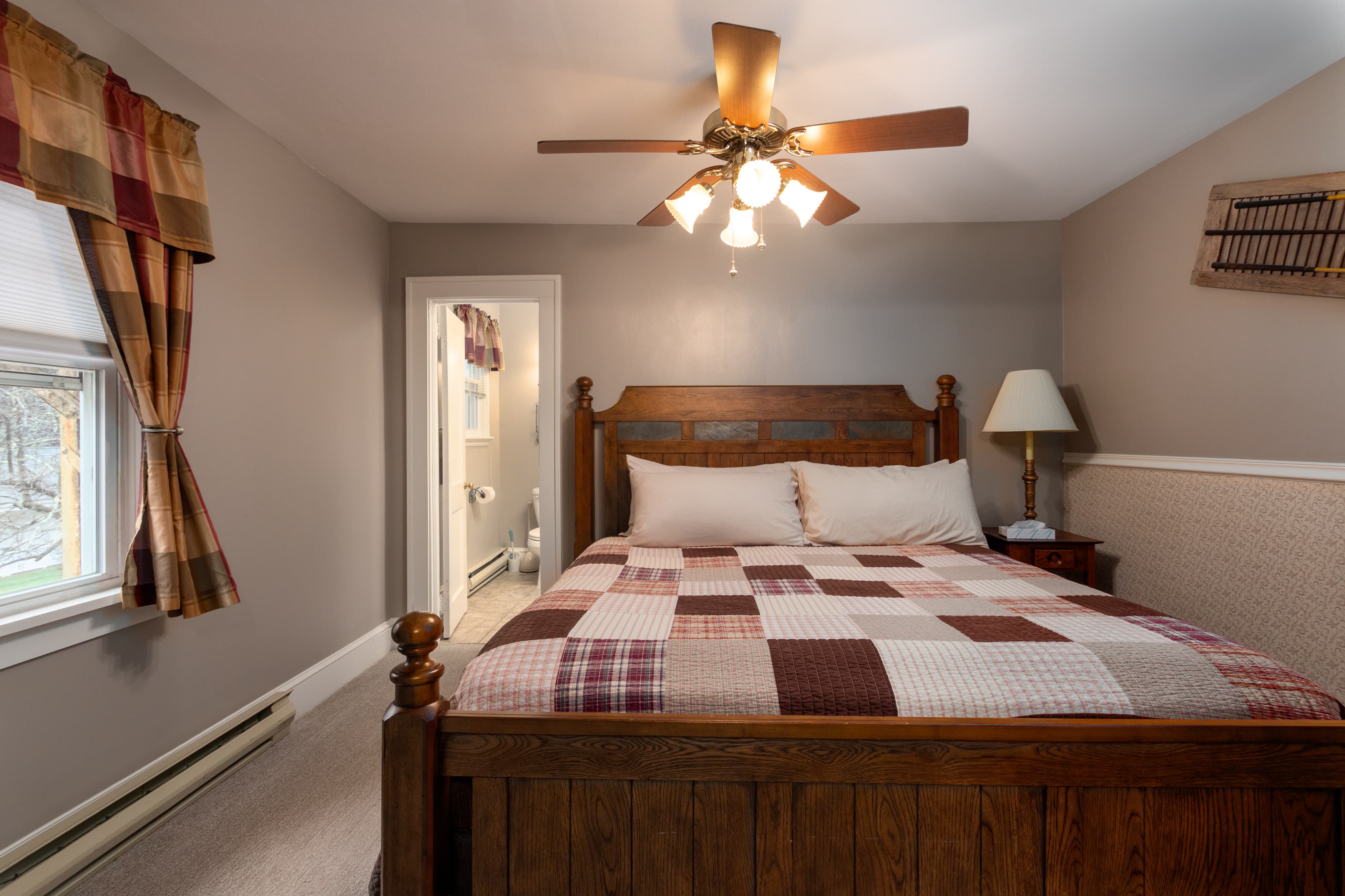A light-filled bedroom with a dark wood king bed, a red and brown patchwork quilt, a window with plaid curtains, and a view into the bathroom.