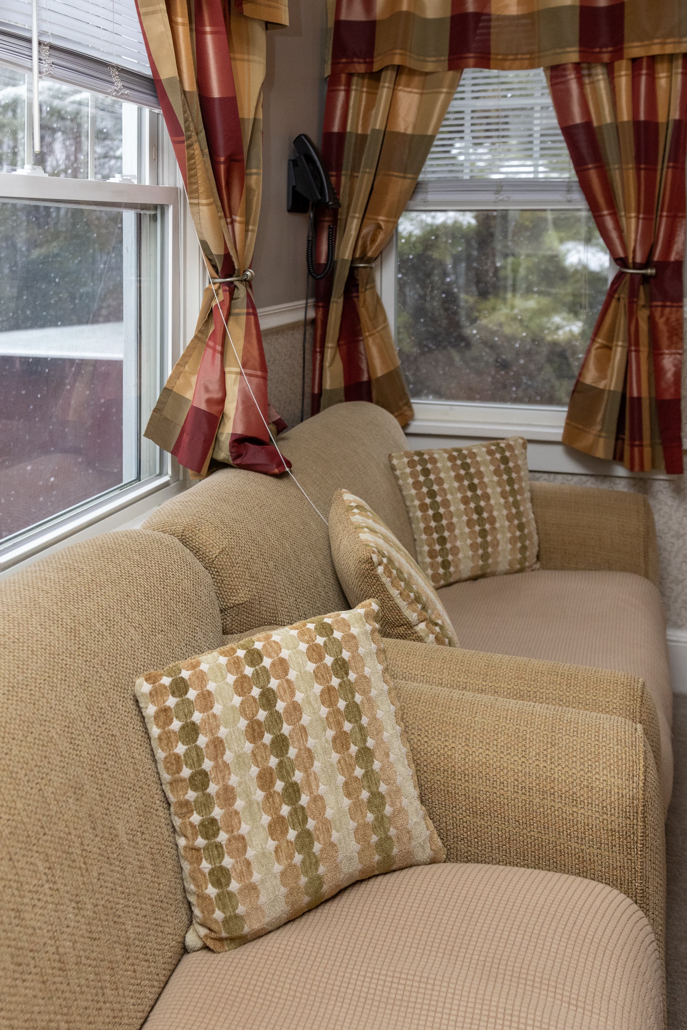 An inviting sunlit sitting area with a tan sofa and patterned pillows, framed by windows with plaid red and gold curtains.