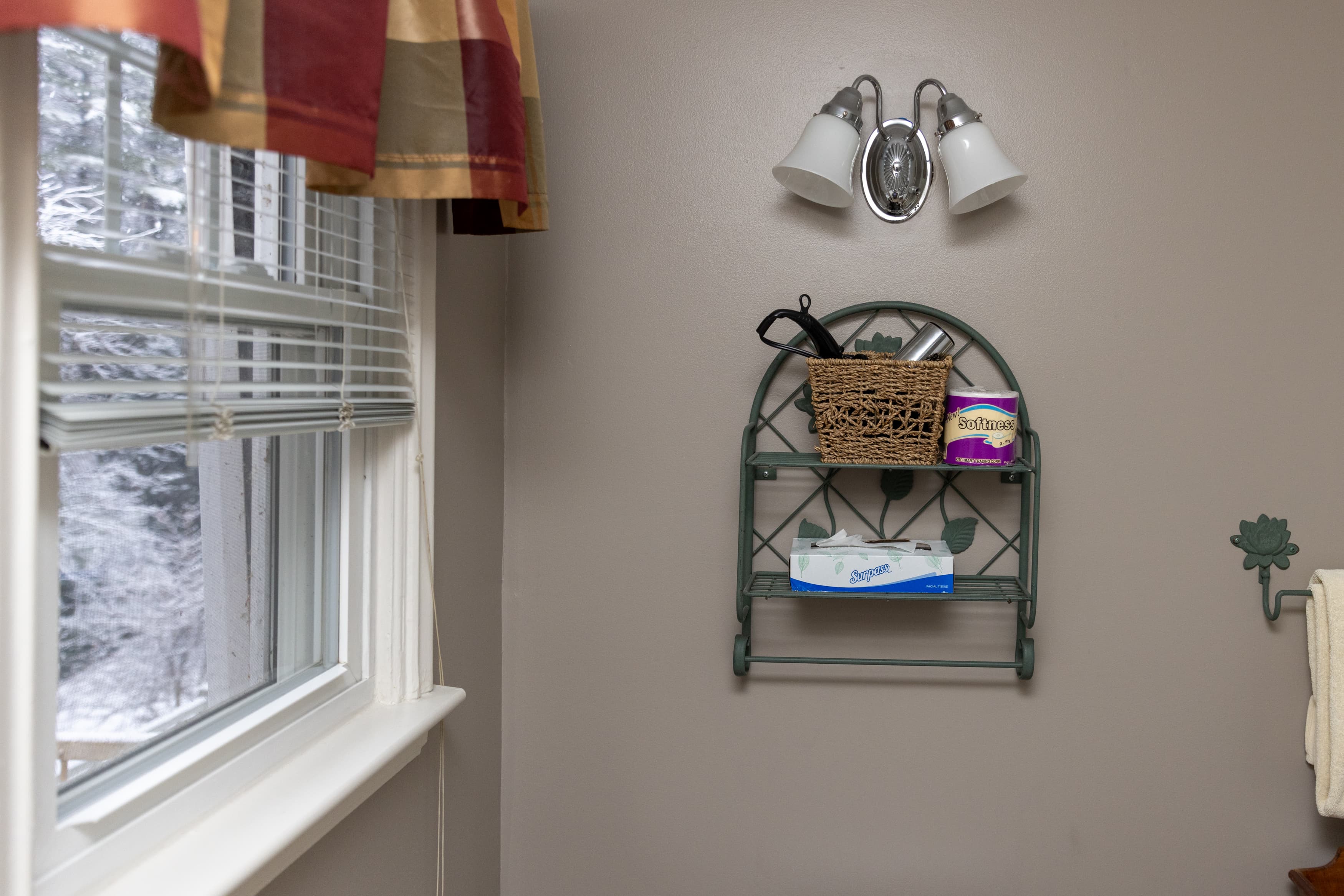A bathroom detail shot featuring a window with plaid valance, a decorative metal shelf holding toiletries, and a three-bulb vanity light.