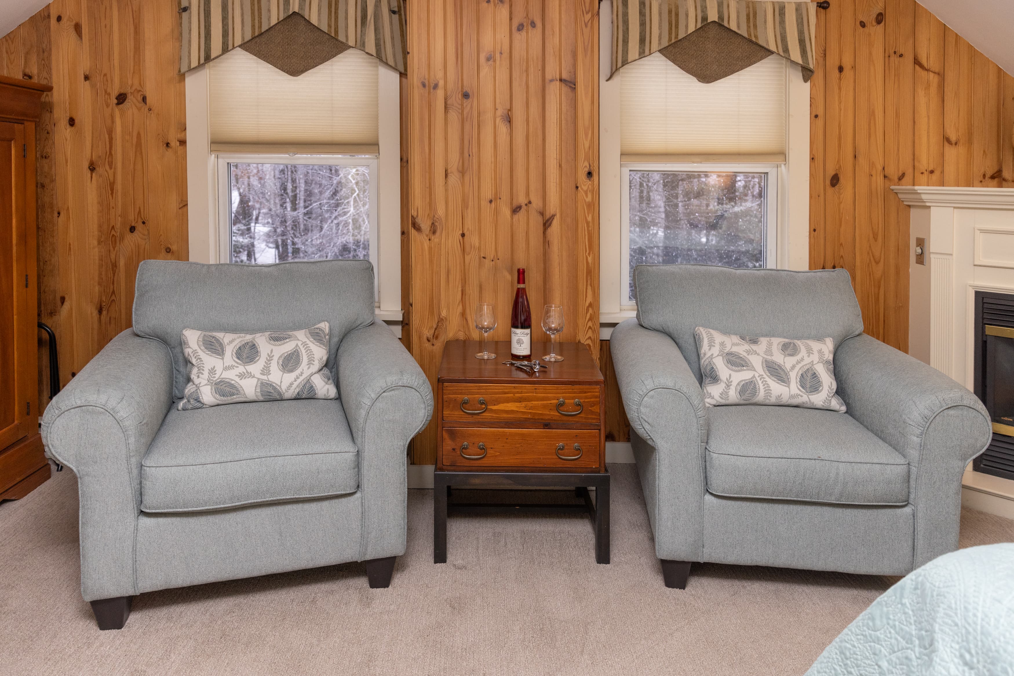 A cozy sitting area in an attic room featuring two light blue armchairs, a small chest nightstand, and a knotty pine paneled wall between two windows.
