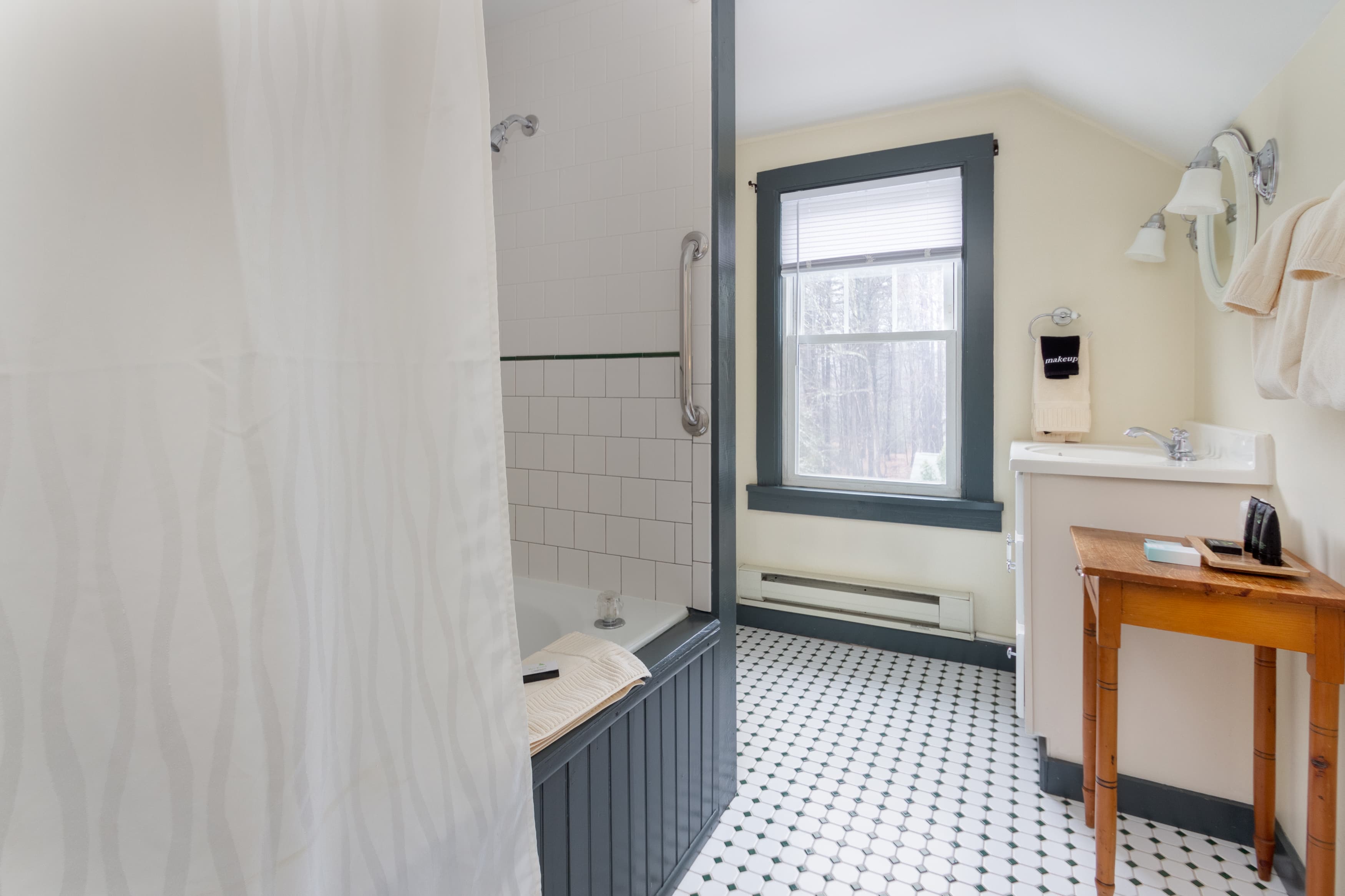 A bright bathroom with a black and white tiled floor, a tub/shower combo with subway tile, and a vanity area with a wooden side table.