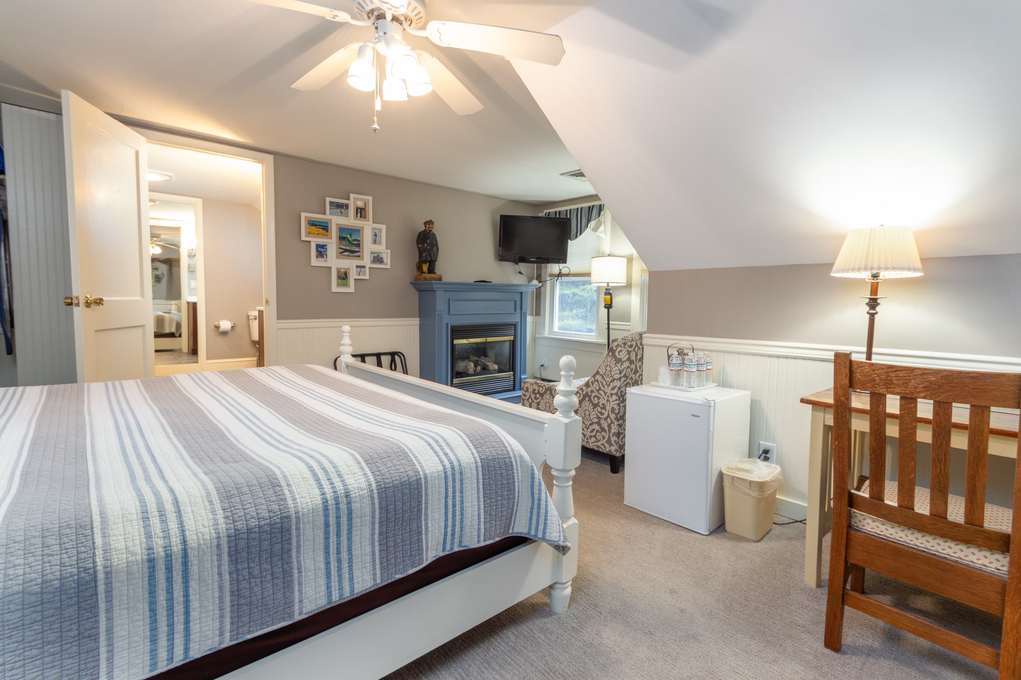 A light-filled third-floor bedroom with sloping ceilings, a king bed with a blue striped quilt, a blue fireplace mantle, a mini-fridge, and a desk.