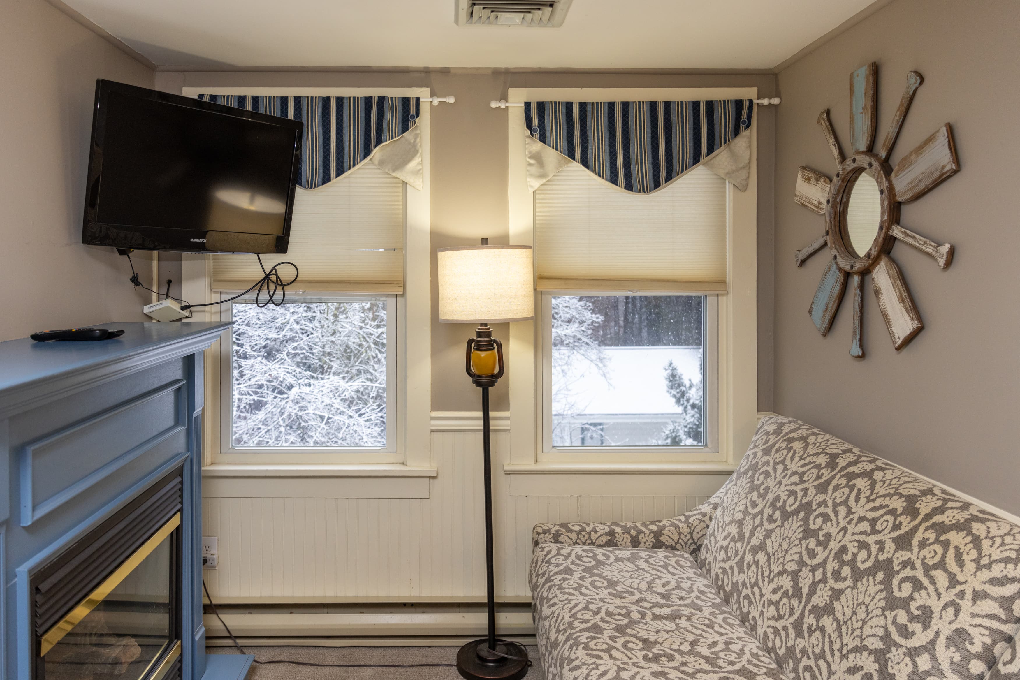 A comfy sitting area with a blue fireplace, a wall-mounted TV, a patterned loveseat, and two windows with blue striped valances.