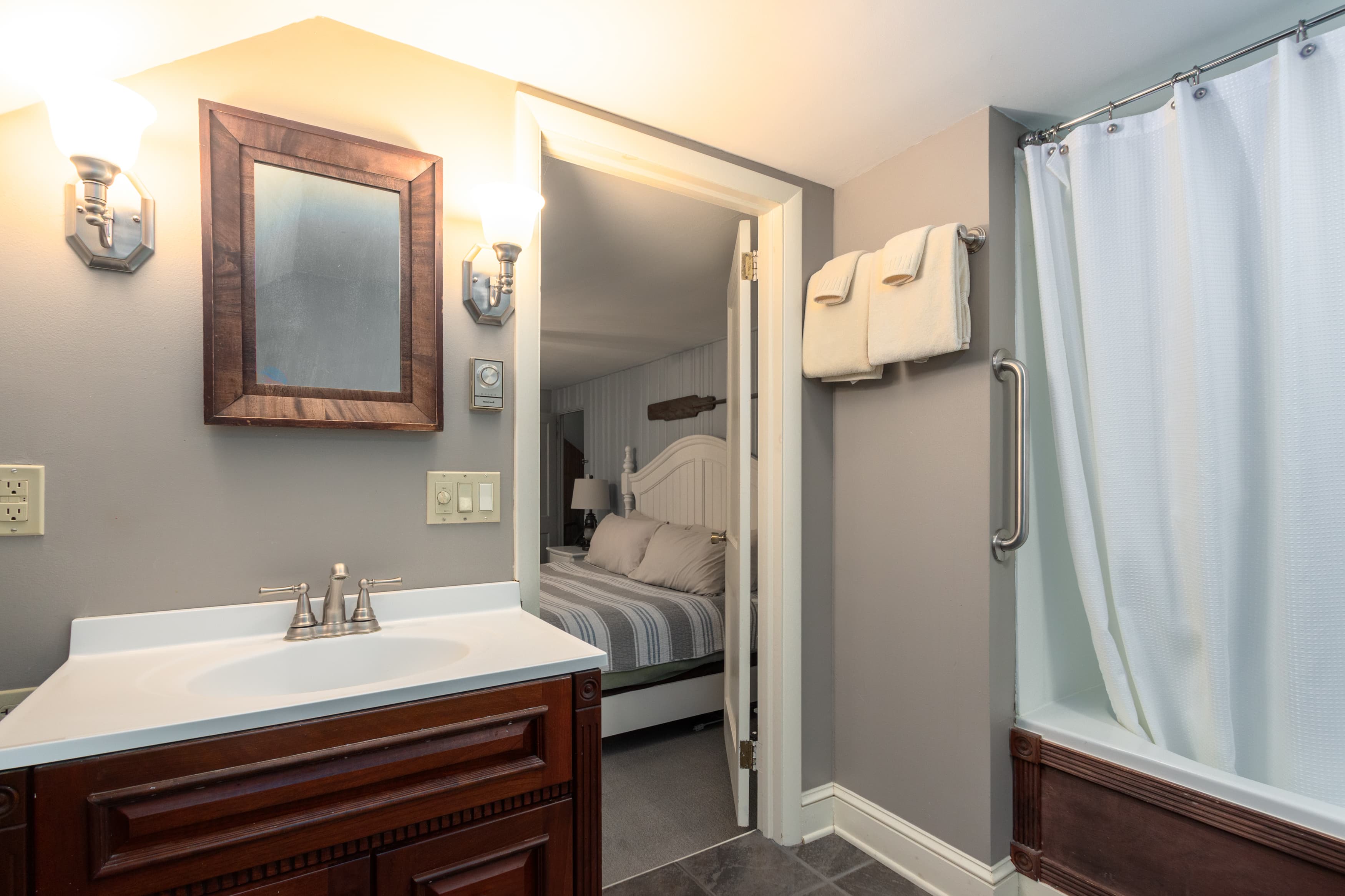 A bathroom with a dark wood vanity, a framed mirror, and a shower, with a view through the doorway into a bedroom with a white headboard.