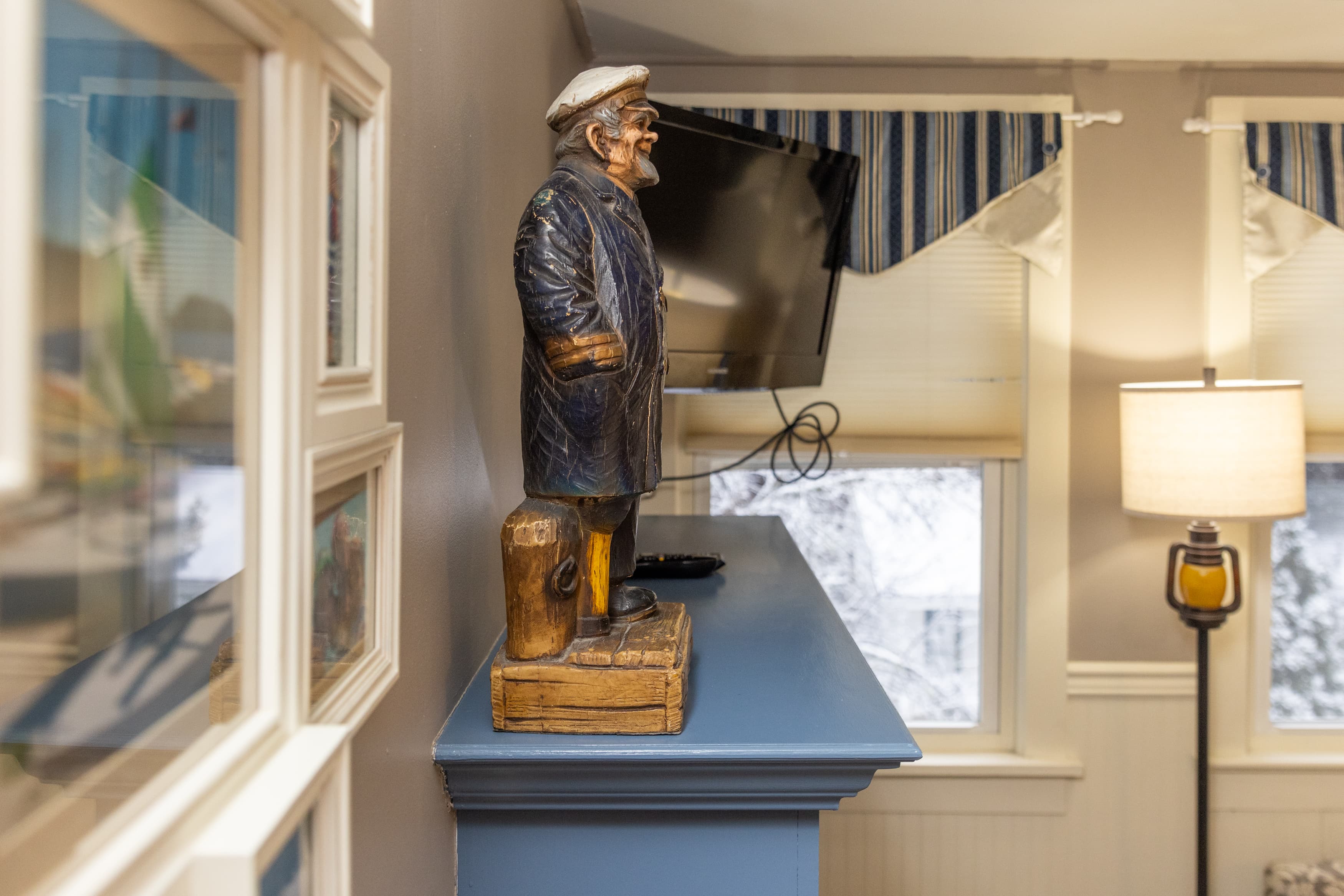 A maritime-themed folk art-style statue of a sailor stands atop a light blue mantel below a wall-mounted TV, next to a wall of framed artwork.