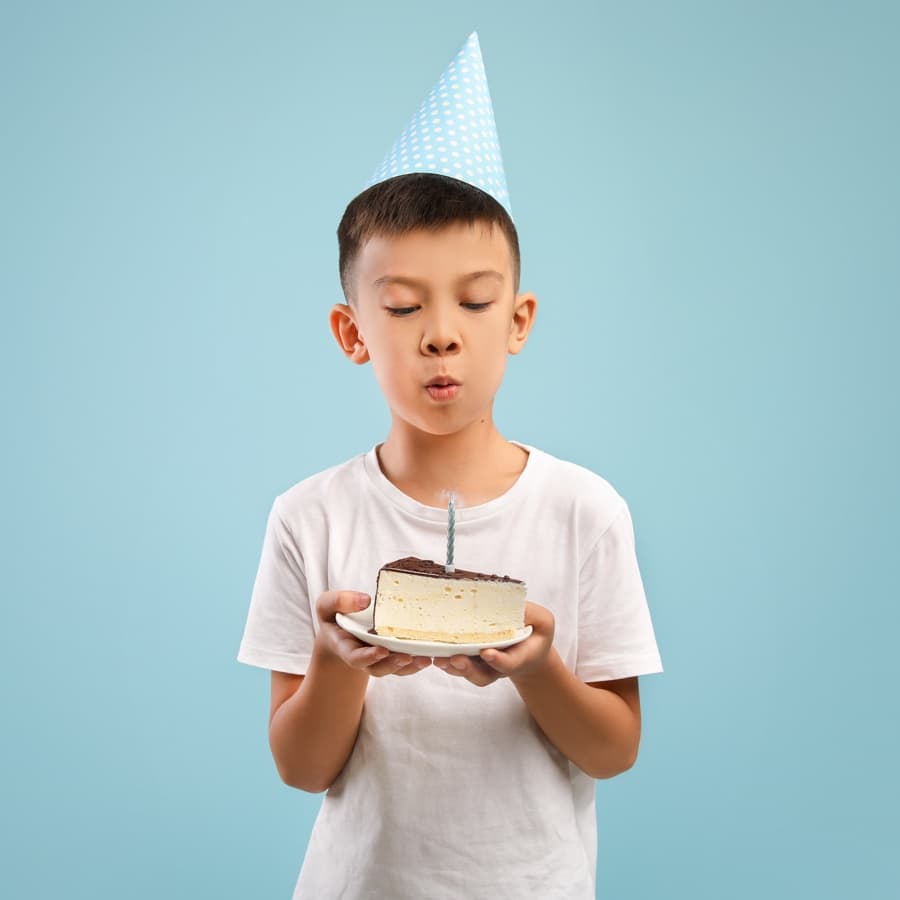 A boy in a party hat blows out a candle on a slice of cake.