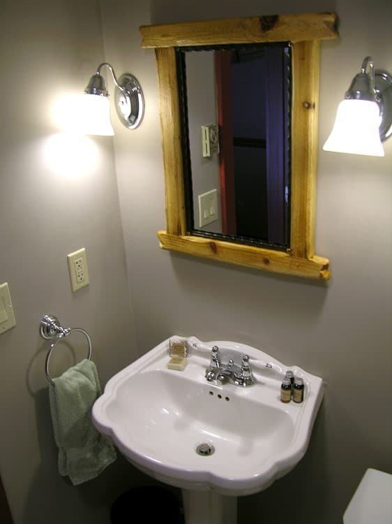 A close-up of a rustic-style bathroom with a white pedestal sink, a wooden-framed mirror, and a pair of sconce lights on the gray wall.