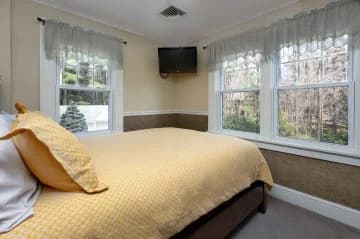 A cozy bedroom featuring a yellow quilted bedspread, two windows with sheer curtains, and a wall-mounted TV.