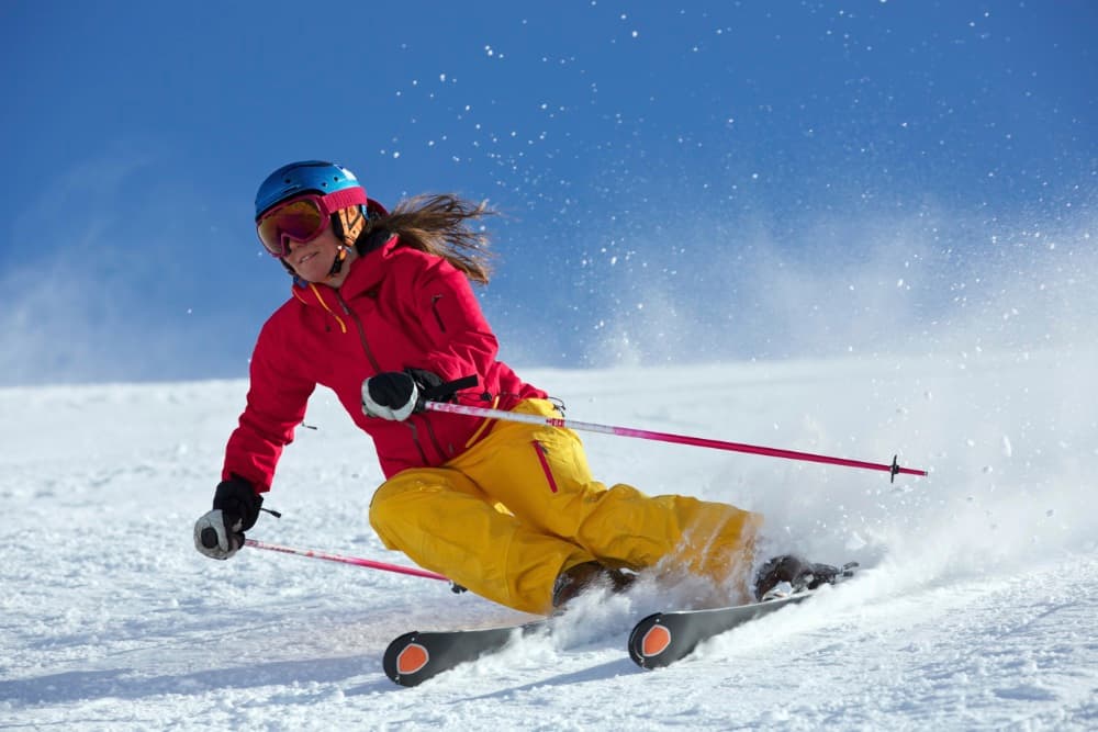 A skier in a red jacket and yellow pants navigates a snowy slope, creating a spray of snow.