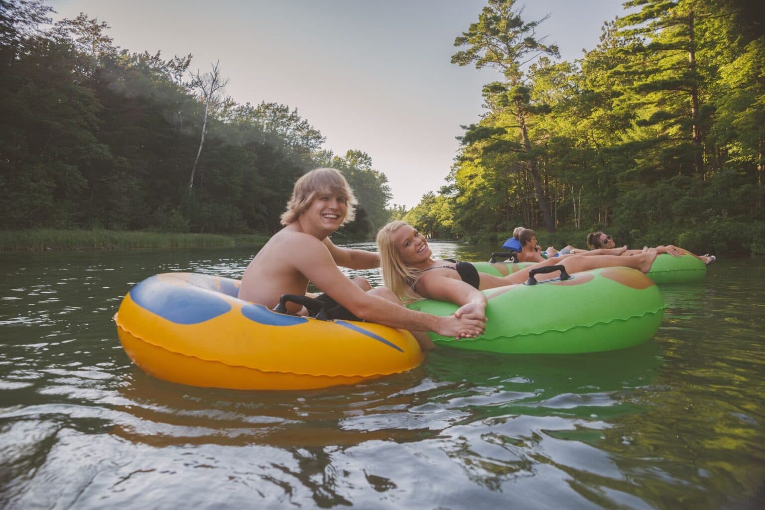 Two friends smile while floating on colorful inner tubes in a serene river surrounded by trees.