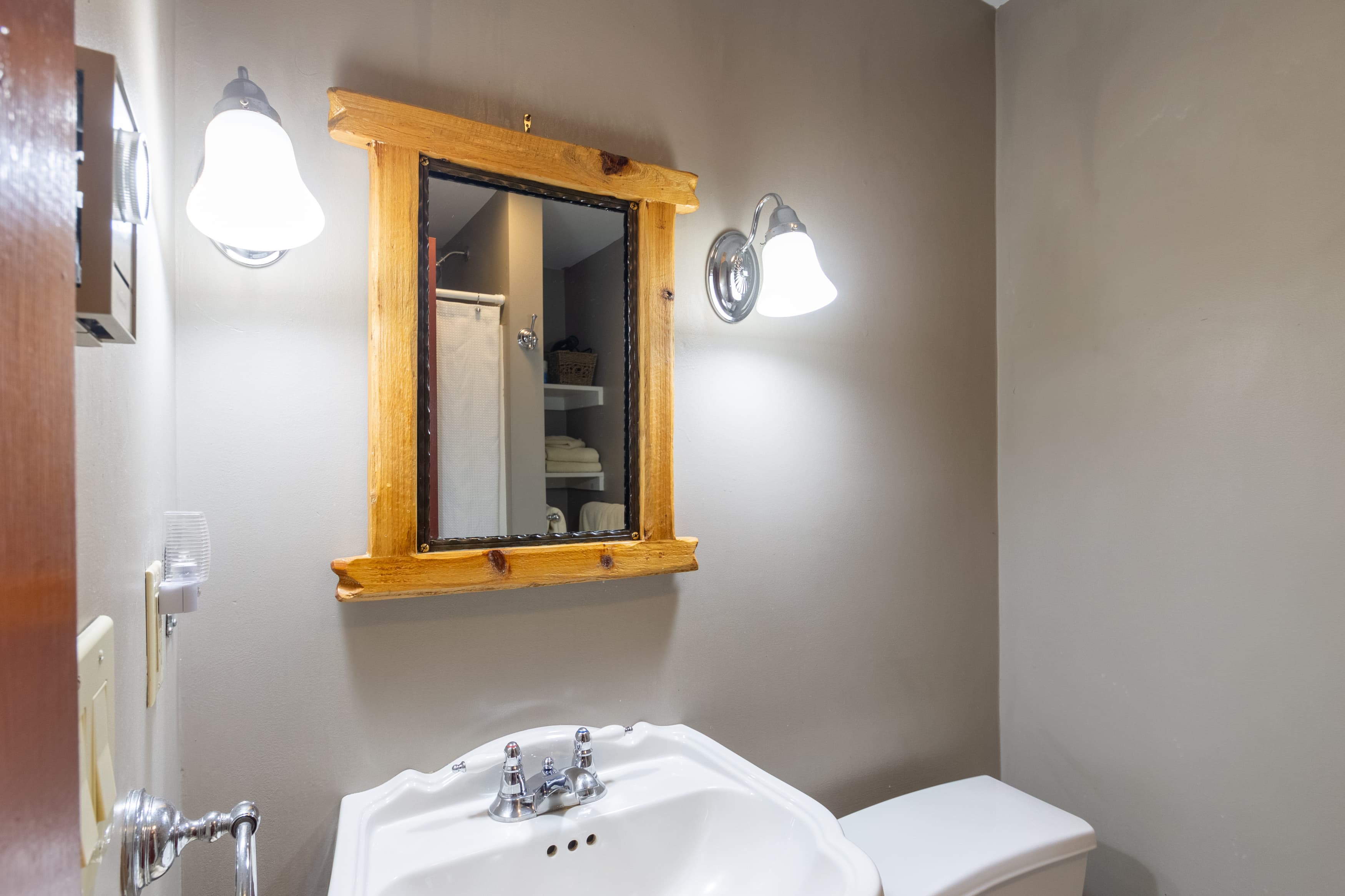 A close-up of a rustic-style bathroom with a white pedestal sink, a wooden-framed mirror, and a pair of sconce lights on the gray wall.