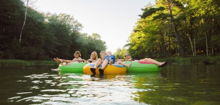Five people relax on colorful inner tubes floating in a tranquil river surrounded by green trees.