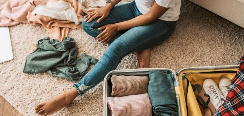 A person sits on the floor surrounded by folded and unfolded clothes near an open suitcase.