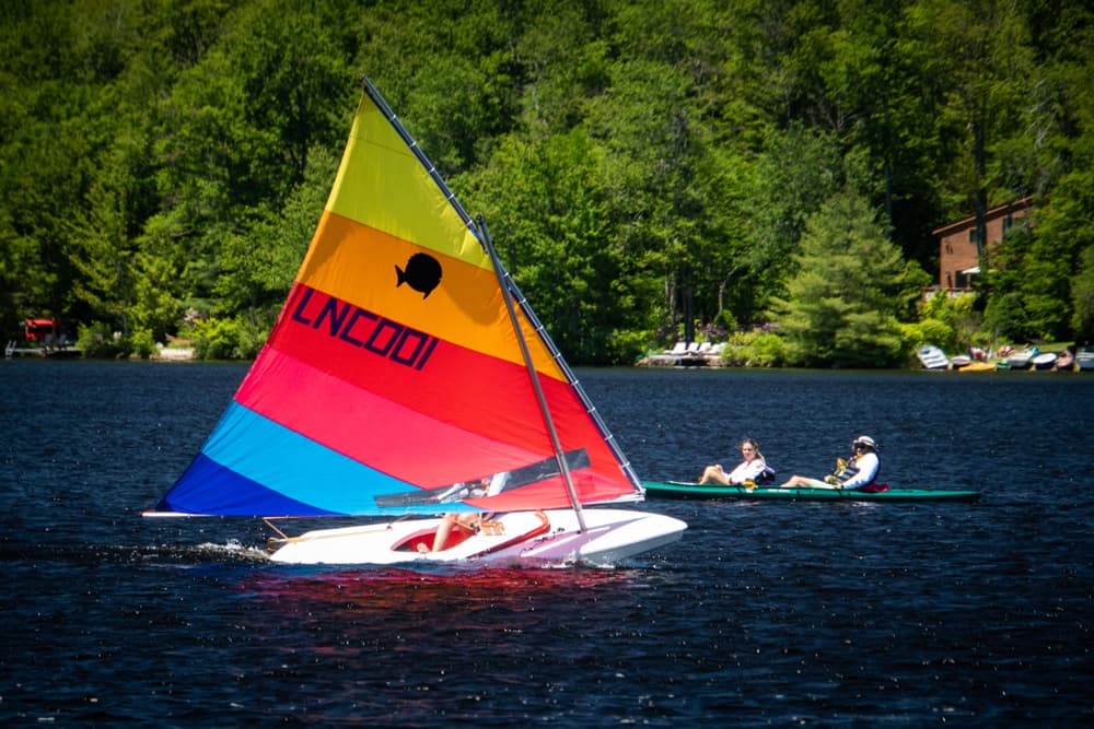 A colorful sailboat glides across a lake with two people kayaking nearby.