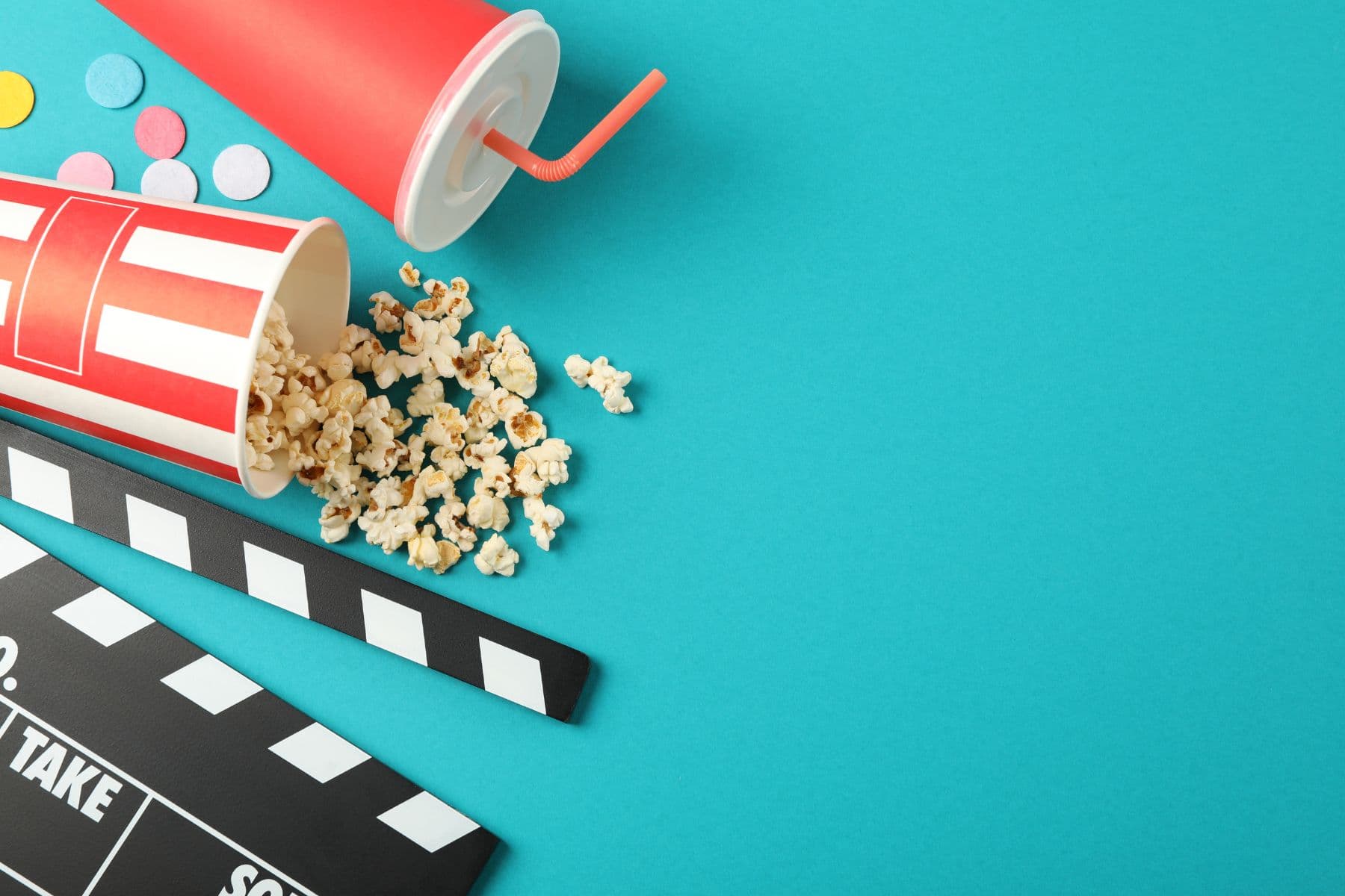 A spilled popcorn container and a drink cup are placed beside a movie clapper on a bright turquoise background.
