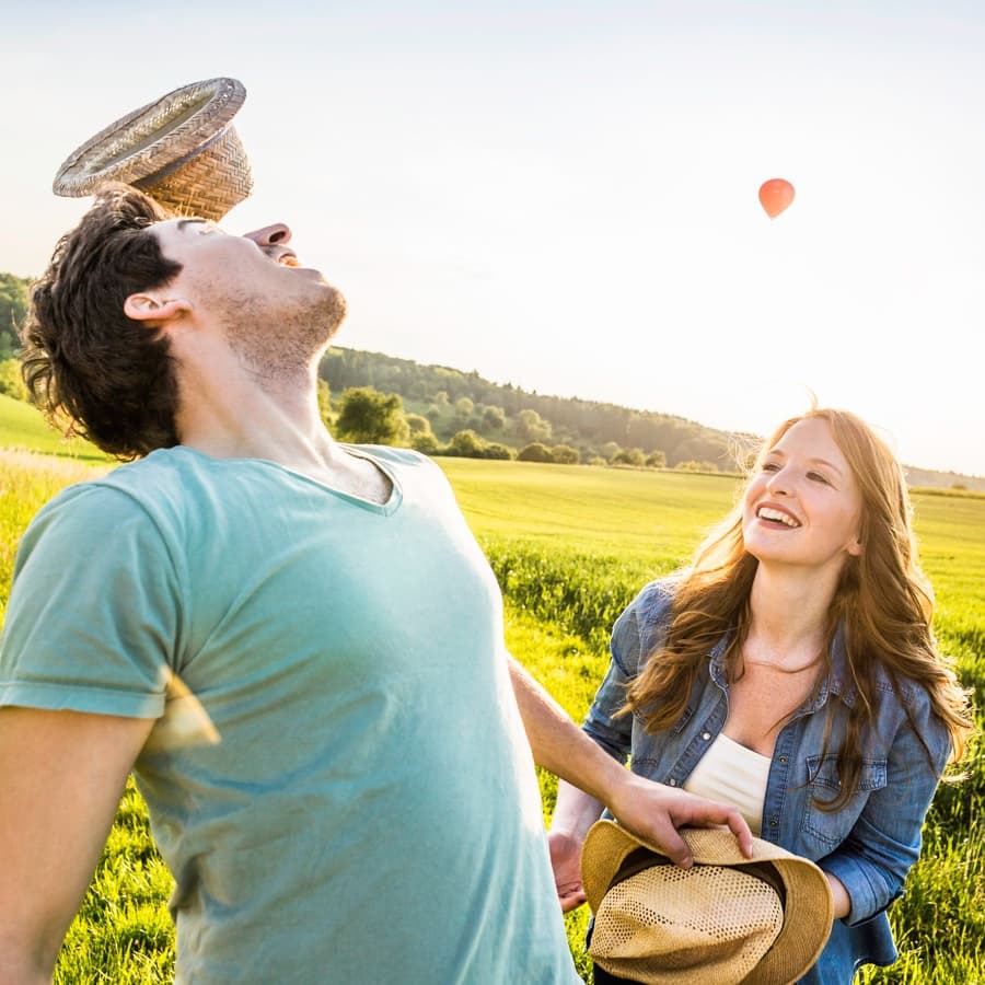 A man balances a straw basket on his head while a woman smiles beside him in a sunny field.