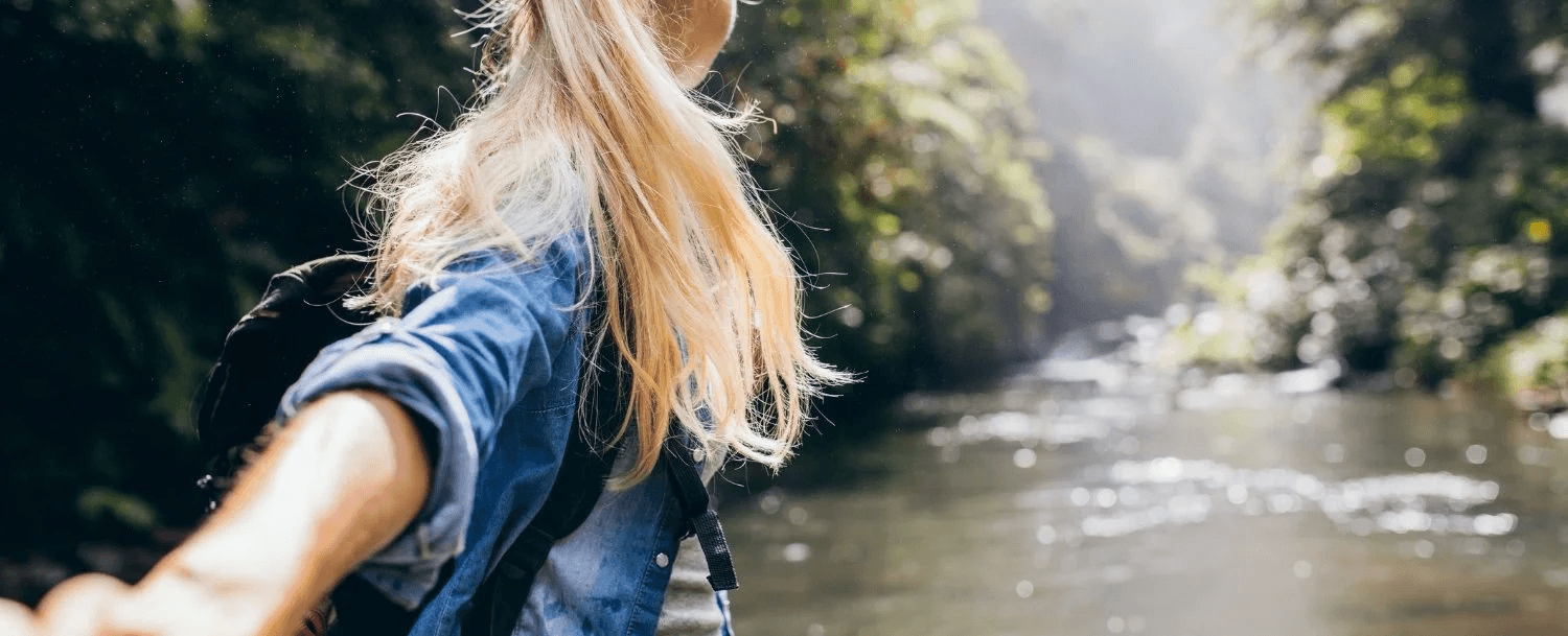 A person with long hair is holding out their hand towards a river surrounded by lush greenery.