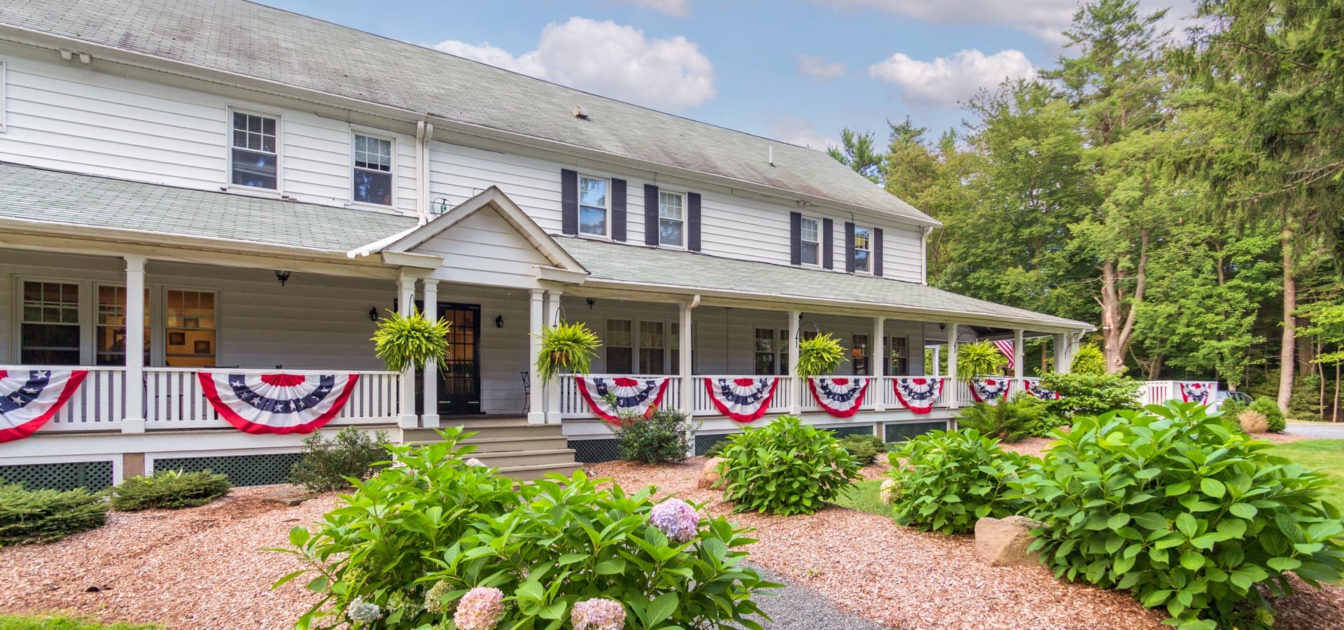 A charming two-story white house with a wraparound porch, adorned with red, white, and blue bunting, surrounded by lush greenery.