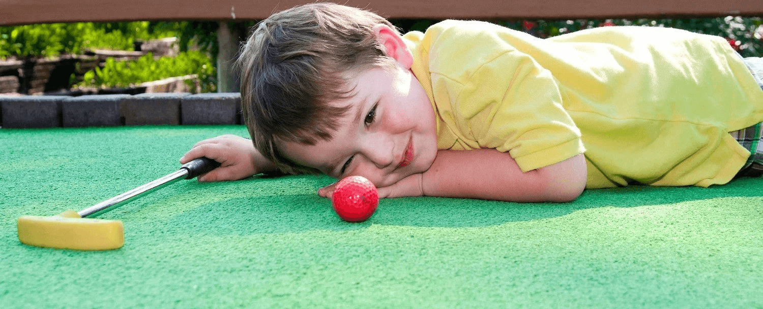 A young boy in a yellow shirt lies on a green mini-golf course, closely examining a red ball with a putter nearby. A young boy in a yellow shirt lies on a green mini-golf course, closely examining a red ball with a putter nearby.