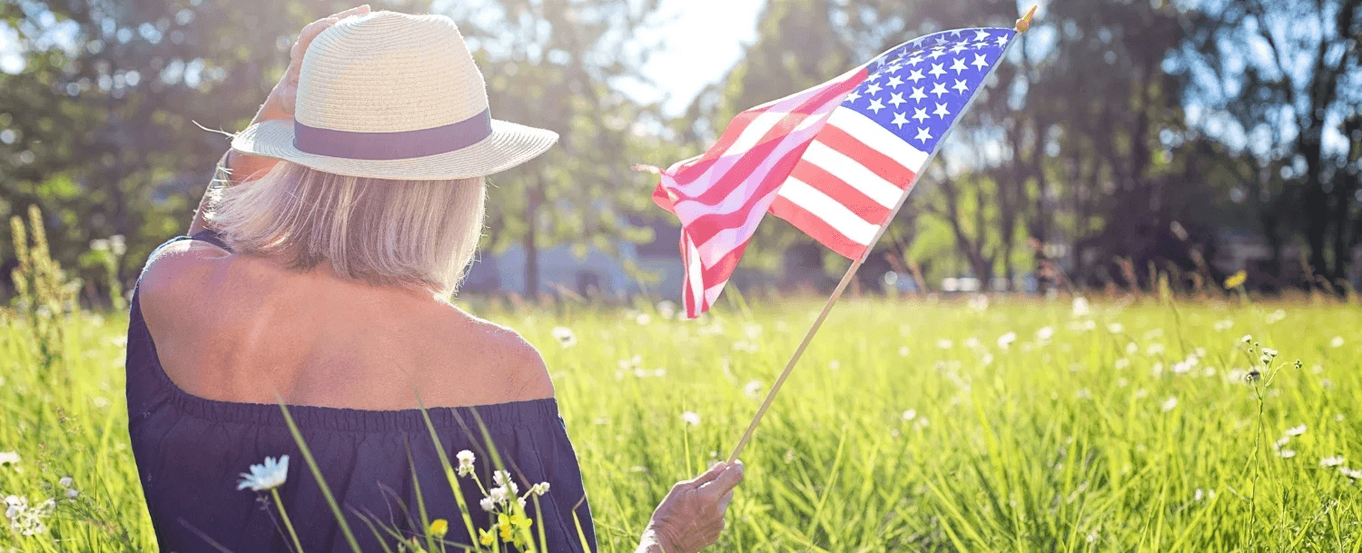 A woman in a straw hat waves an American flag while standing in a sunny field.