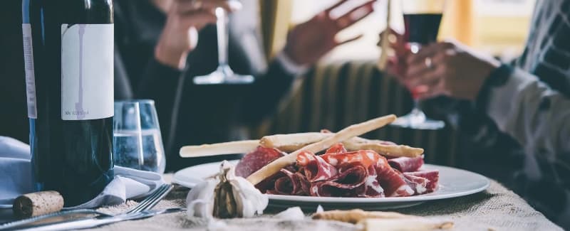 A table set with wine, glasses, and a plate of cured meats and breadsticks, with hands gesturing in conversation.
