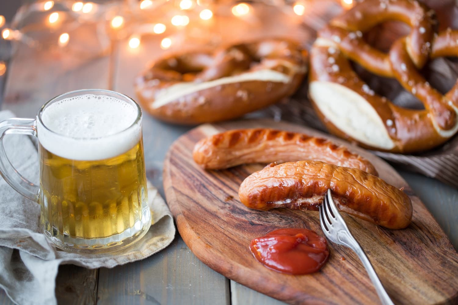 A wooden board with a sausage, fork, ketchup, pretzels, and a mug of beer, set against a backdrop of warm lights. A wooden board with a sausage, fork, ketchup, pretzels, and a mug of beer, set against a backdrop of warm lights.