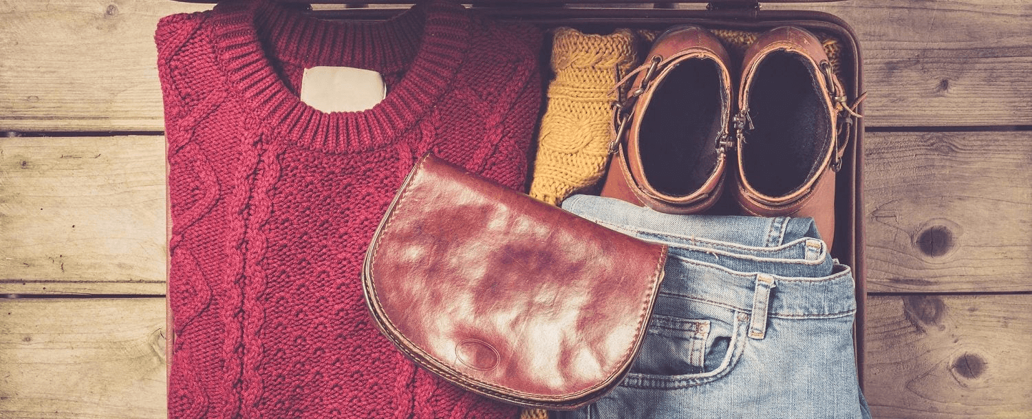 A suitcase filled with a red sweater, yellow knitwear, denim jeans, brown shoes, and a brown leather bag.
