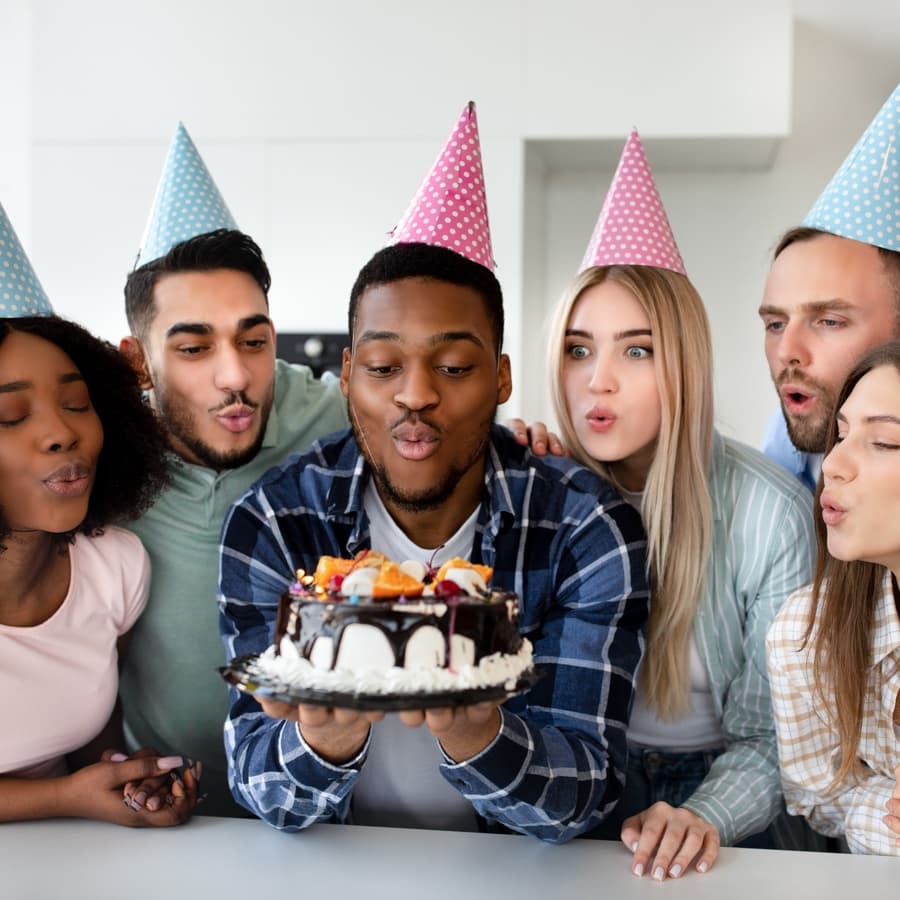 A group of six friends wearing party hats gather around a cake, preparing to blow out the candles.