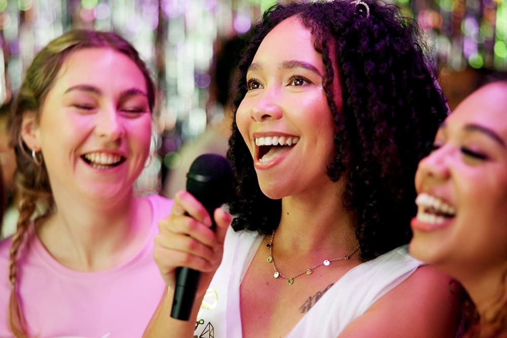 Three friends joyfully sing together with a microphone in a festive setting.