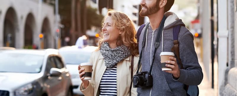 A smiling couple walks together on a city street, each holding a coffee cup.