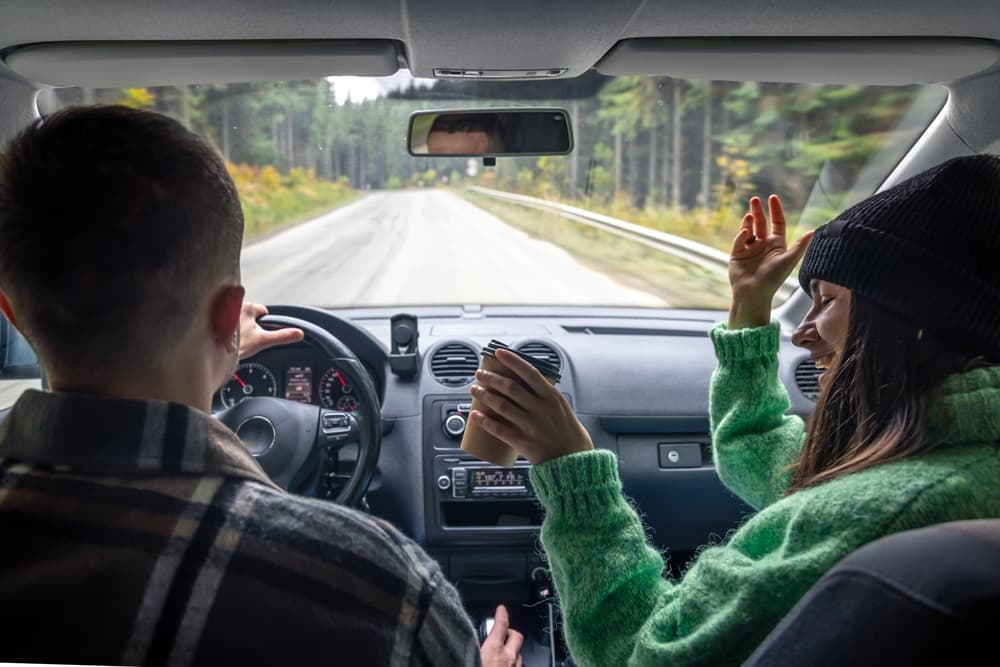 A person drives while another passenger joyfully gestures and holds a drink in a car.