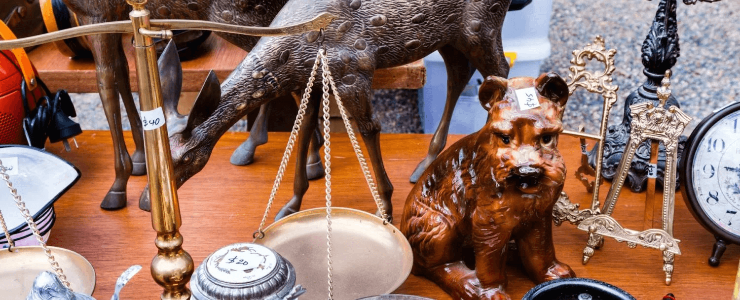 A wooden table displays various antique items, including a scale, metal containers, a deer figurine, and a dog statue.