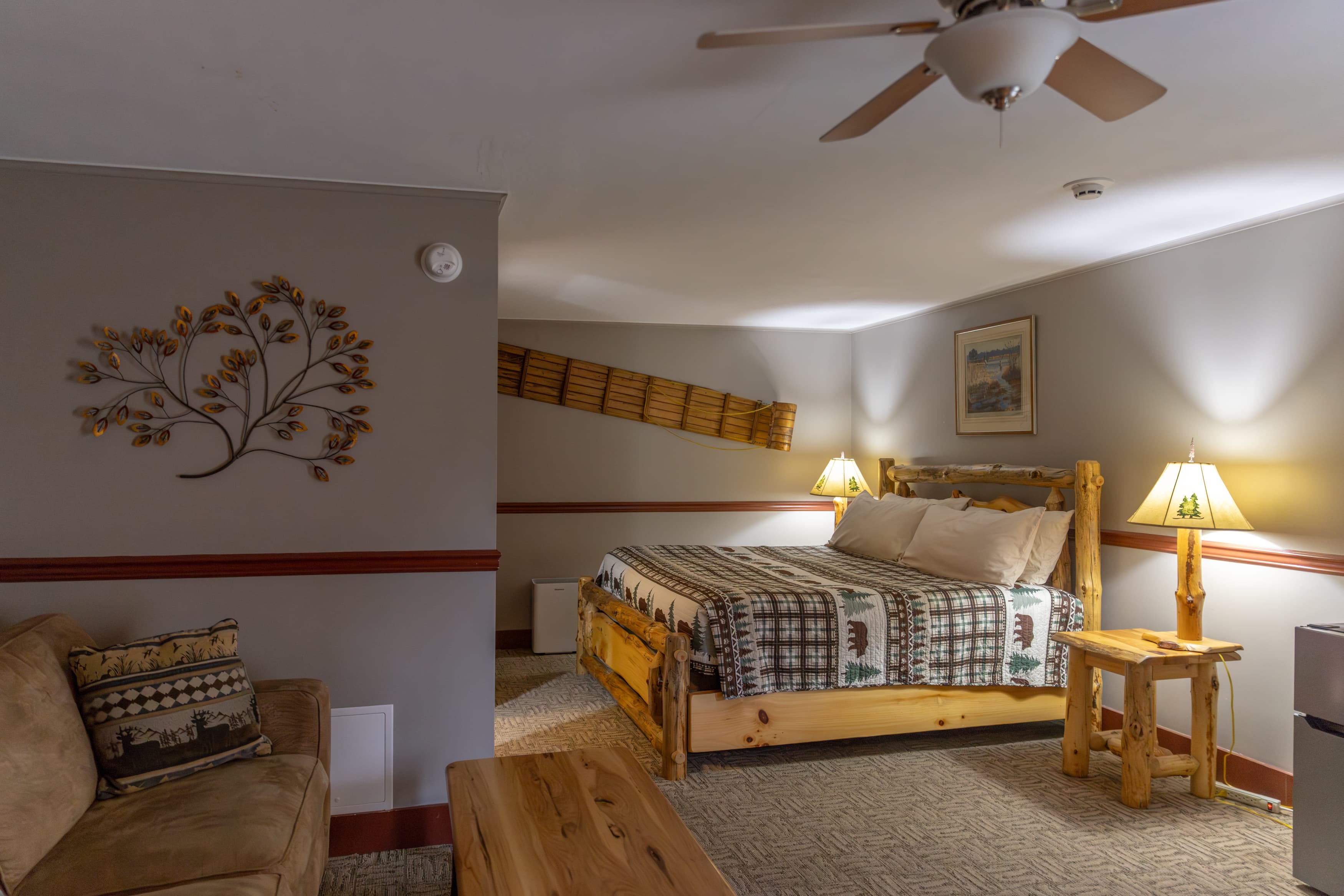 An interior view of a rustic, cabin-themed suite featuring a log-style queen bed and a tan sofa in a cozy, carpeted room.