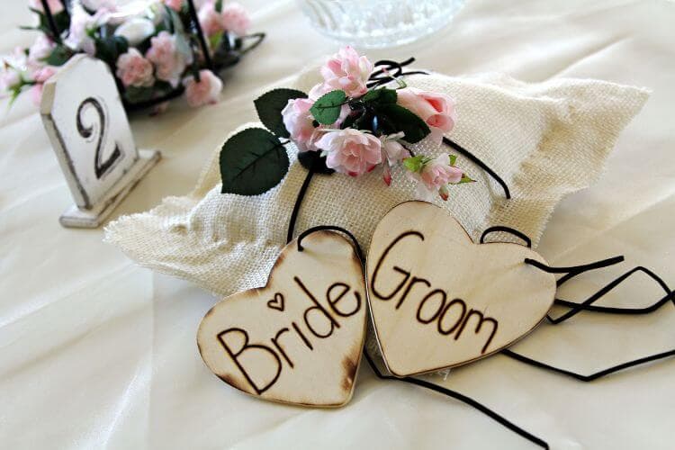 Wooden heart decorations labeled "Bride" and "Groom" alongside a small bouquet on a decorative pillow.