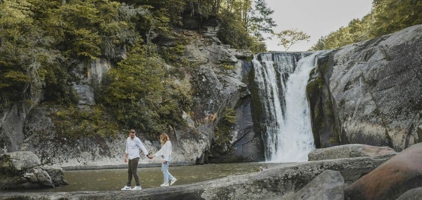 A couple holds hands while walking on rocks near a waterfall surrounded by greenery.