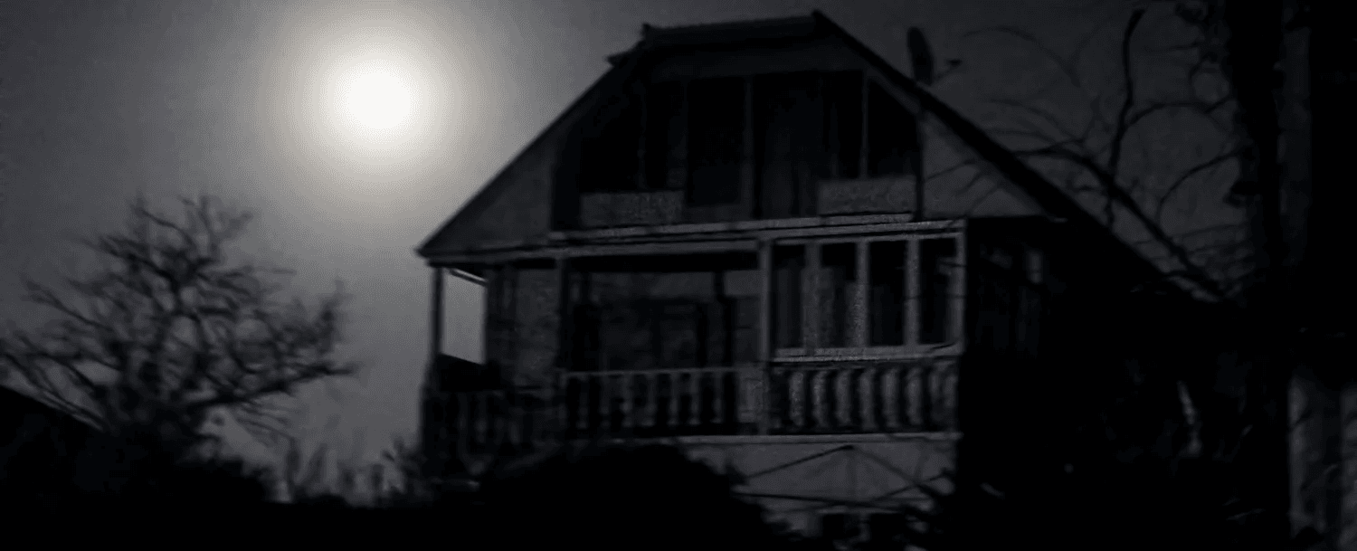 A spooky, dimly lit house silhouetted against a full moon in a dark sky.