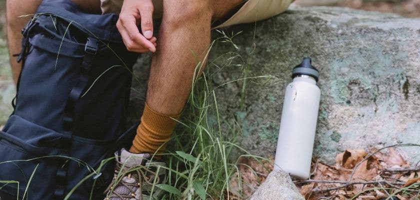 A person sits beside a backpack and a water bottle on a stone in a grassy outdoor setting.