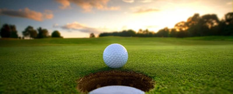 A close-up of a golf ball poised near a hole on a green, with a sunset in the background.