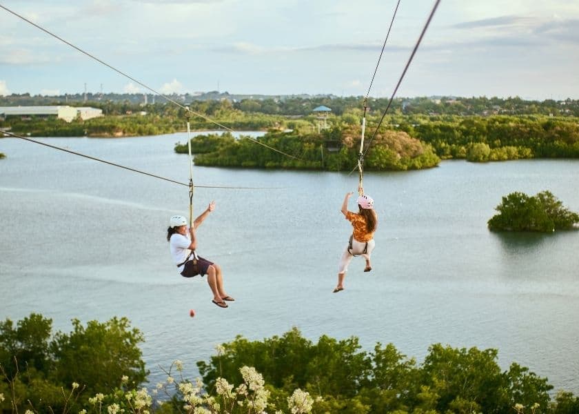 Two individuals ziplining over a body of water surrounded by greenery. Two individuals ziplining over a body of water surrounded by greenery.