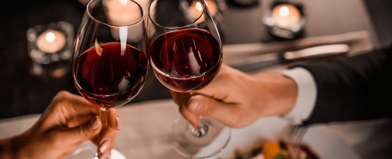 Two hands clink wine glasses in a softly lit dinner setting.